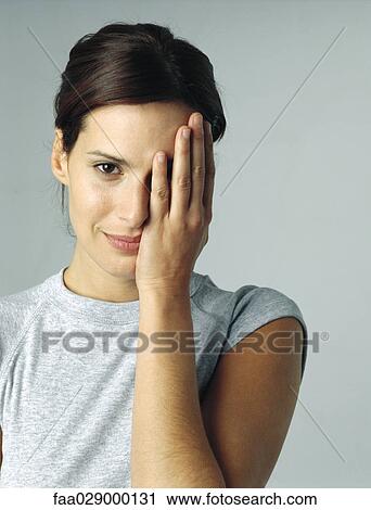Stock Image - Woman covering one eye with hand, smiling at camera, portrait. Fotosearch