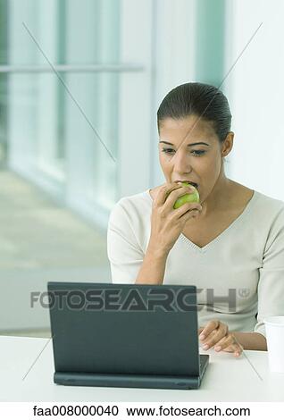 Stock Image - Woman eating apple and using laptop. Fotosearch