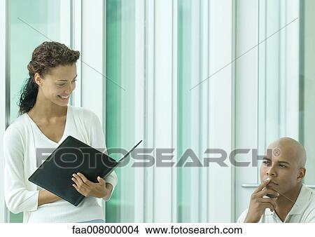 Woman holding agenda smiling while male colleague thinks View Large Photo Image Picture - Woman holding agenda smiling while male colleague thinks. Fotosearch