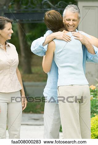 Stock Image - Woman hugging man as second woman watches smiling. Fotosearch
