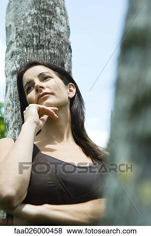 Stock Photo - Woman leaning against tree trunk, hand under chin, low angle view. Fotosearch