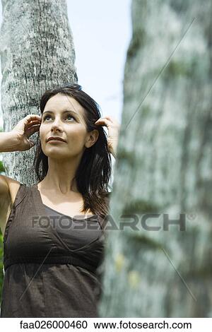Woman leaning against tree trunk, pushing hair behind ears, looking up View Large Photo Image Stock Image - Woman leaning against tree trunk, pushing hair behind ears, looking up. Fotosearch