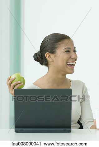 Woman sitting at laptop, holding up apple, laughing View Large Photo Image Stock Image - Woman sitting at laptop, holding up apple, laughing. Fotosearch