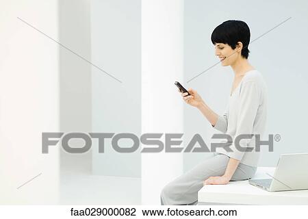 Woman sitting on edge of desk, looking at cell phone and smiling, side view View Large Photo Image Stock Image - Woman sitting on edge of desk, looking at cell phone and smiling, side view. Fotosearch
