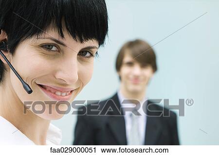 Woman wearing headset, smiling at camera, businessman in background View Large Photo Image Stock Image - Woman wearing headset, smiling at camera, businessman in background. Fotosearch