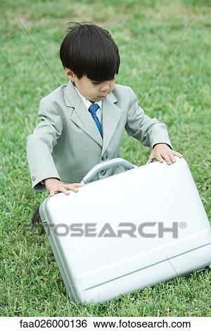 Stock Photograph - Young boy dressed in suit, crouching in grass, opening briefcase. Fotosearch