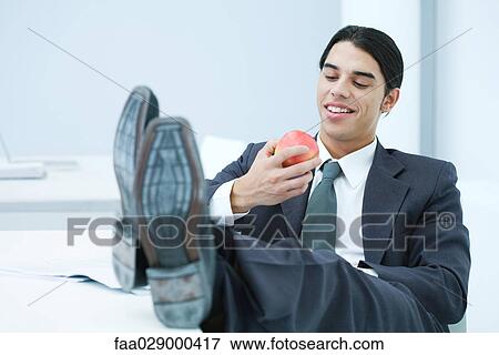 Stock Photo - Young businessman sitting at desk with feet up, eating apple. Fotosearch