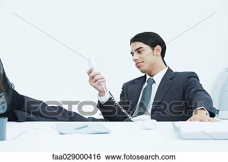 Young businessman sitting at desk with feet up, looking at landline phone, smiling View Large Photo Image Stock Photograph - Young businessman sitting at desk with feet up, looking at landline phone, smiling. Fotosearch