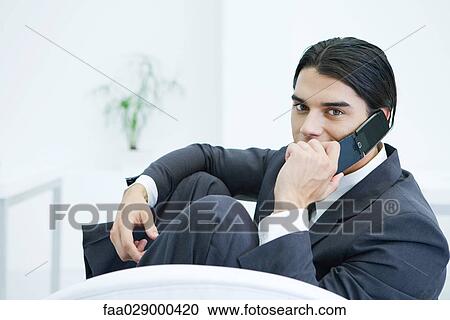 Young businessman sitting with knees up, using cell phone, looking at camera View Large Photo Image Stock Image - Young businessman sitting with knees up, using cell phone, looking at camera. Fotosearch