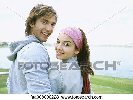 Young couple standing by lake, smiling over shoulder at camera View Large Photo Image Stock Photo - Young couple standing by lake, smiling over shoulder at camera. Fotosearch