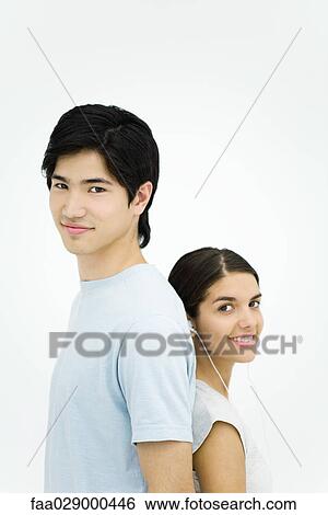 Stock Photograph - Young man and woman standing back to back, smiling at camera, portrait. Fotosearch