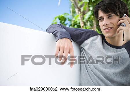 Stock Image - Young man leaning against wall, using cell phone. Fotosearch