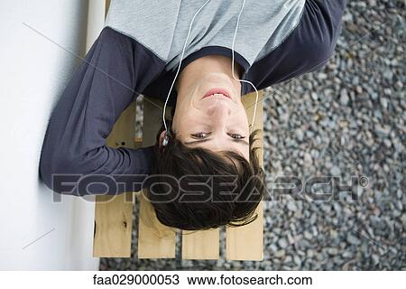 Stock Image - Young man lying on bench, listening to earphones, looking at camera. Fotosearch
