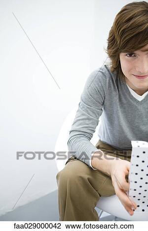 Young man sitting, holding gift bag, smiling at camera, cropped view View Large Photo Image Stock Image - Young man sitting, holding gift bag, smiling at camera, cropped view. Fotosearch