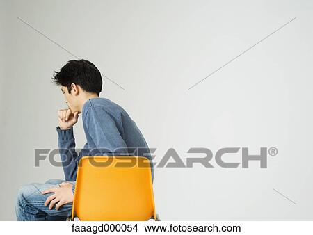 Picture - Young man sitting on chair, holding chin. Fotosearch