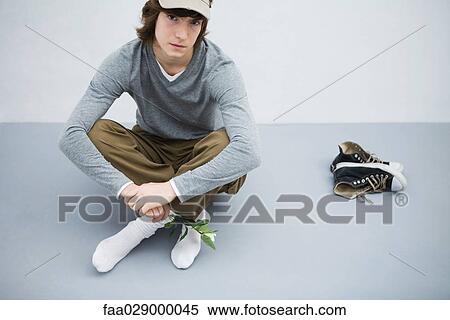 Stock Photography - Young man sitting on the ground next to his shoes, holding flower, looking at camera. Fotosearch