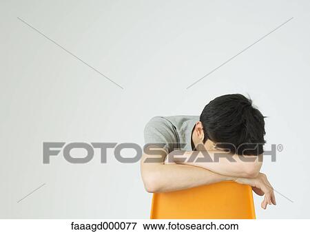 Stock Photo - Young man sitting with head down on back of chair. Fotosearch