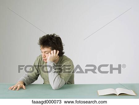 Stock Image - Young man talking on phone, turning away from open notebook on table. Fotosearch
