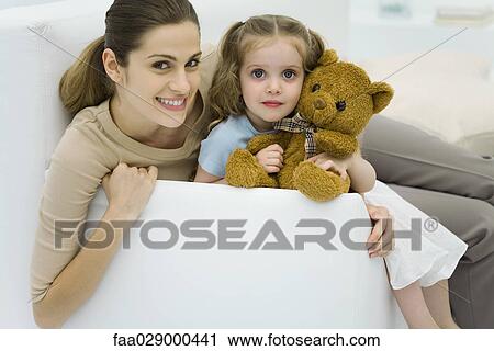 Stock Image - Young mother sitting with daughter on sofa, little girl holding teddy bear, smiling at camera. Fotosearch