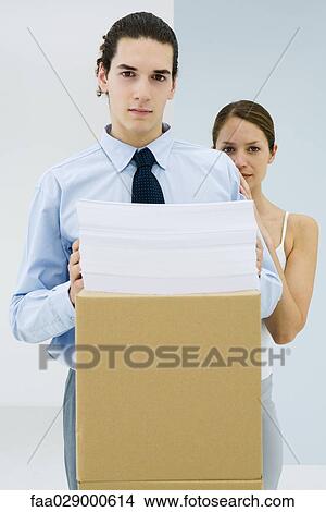 Picture - Young office workers, young man holding stack of paper on cardboard box, looking at camera. Fotosearch