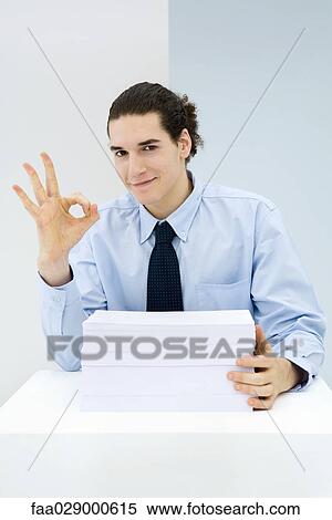 Young professional sitting behind stack of papers, making OK hand gesture, portrait View Large Photo Image Stock Photography - Young professional sitting behind stack of papers, making OK hand gesture, portrait. Fotosearch