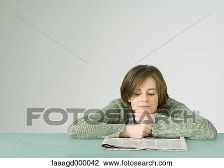 Stock Image - Young woman leaning on table, reading newspaper. Fotosearch