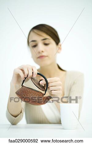 Stock Image - Young woman looking at teapot, lifting lid, focus on foreground. Fotosearch