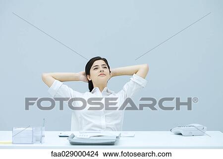 Young woman sitting at desk with hands behind head, looking away, smiling View Large Photo Image Picture - Young woman sitting at desk with hands behind head, looking away, smiling. Fotosearch