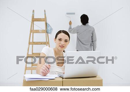 Stock Photo - Young woman sitting at makeshift desk, using laptop computer, smiling at camera, man painting wall in background. Fotosearch