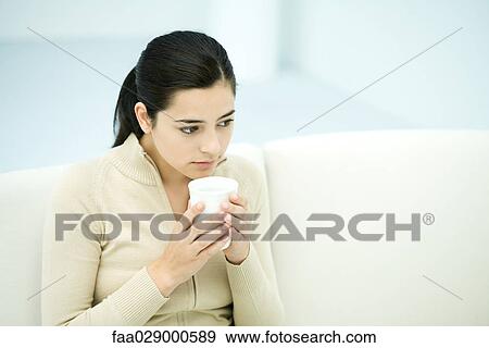 Stock Photo - Young woman sitting, holding coffee mug, looking away. Fotosearch