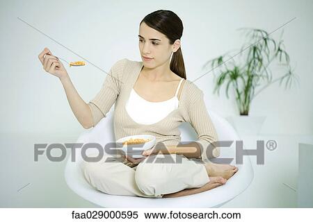 Young woman sitting in chair, holding cereal bowl, looking at spoon View Large Photo Image Stock Photography - Young woman sitting in chair, holding cereal bowl, looking at spoon. Fotosearch