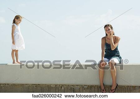 Young woman sitting on low wall, using cell phone, little girl standing nearby, both smiling View Large Photo Image Stock Image - Young woman sitting on low wall, using cell phone, little girl standing nearby, both smiling. Fotosearch