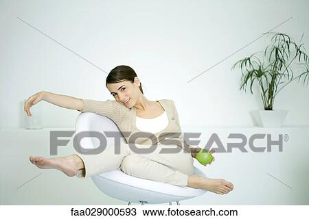 Young woman slouching in chair, holding apple, smiling at camera View Large Photo Image Stock Image - Young woman slouching in chair, holding apple, smiling at camera. Fotosearch