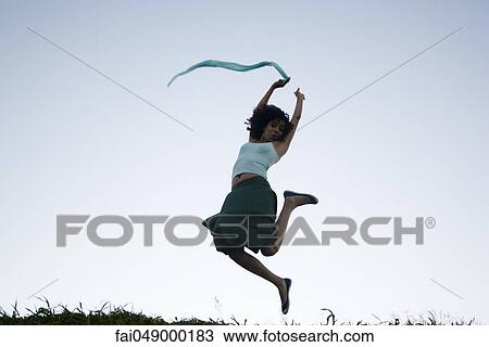 Stock Image - Carefree young woman leaping, scarf in hand trailing behind. Fotosearch