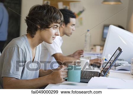 Stock Photo - College student using laptop computer, cup of coffee in hand. Fotosearch