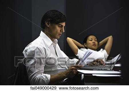 Man in office working late, colleague relaxing in background View Large Photo Image Stock Photo - Man in office working late, colleague relaxing in background. Fotosearch
