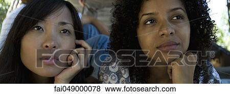 Two young women sitting together outdoors, both looking away, daydreaming View Large Photo Image Stock Photo - Two young women sitting together outdoors, both looking away, daydreaming. Fotosearch