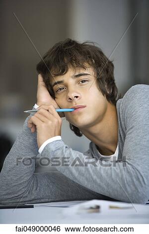 Young man leaning on elbow, pen in mouth, looking at camera with bored expression View Large Photo Image Stock Photograph - Young man leaning on elbow, pen in mouth, looking at camera with bored expression. Fotosearch