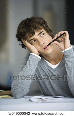 Stock Image - Young man with pen in mouth looking away, daydreaming. Fotosearch