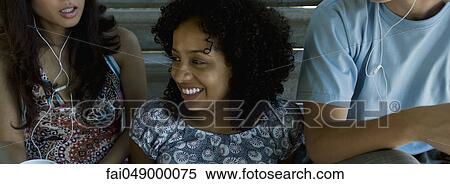 Young woman sitting between two friends listening to earphones, looking away and smiling View Large Photo Image Stock Photography - Young woman sitting between two friends listening to earphones, looking away and smiling. Fotosearch