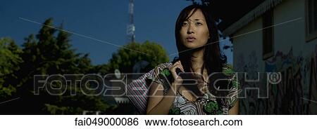 Young woman standing outdoors holding purse over shoulder View Large Photo Image Stock Photograph - Young woman standing outdoors holding purse over shoulder. Fotosearch