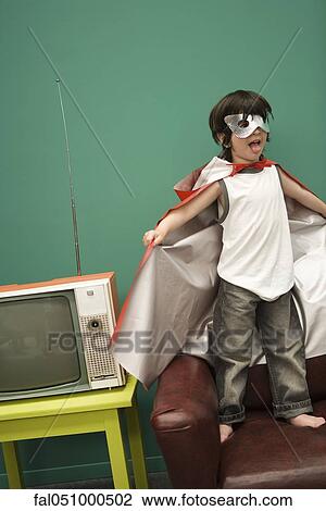 Stock Image - Boy in superhero costume standing on sofa. Fotosearch
