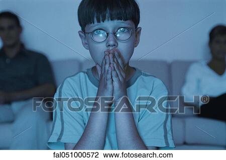 Boy watching TV with hands clasped over mouth, parents sitting in background View Large Photo Image Stock Image - Boy watching TV with hands clasped over mouth, parents sitting in background. Fotosearch