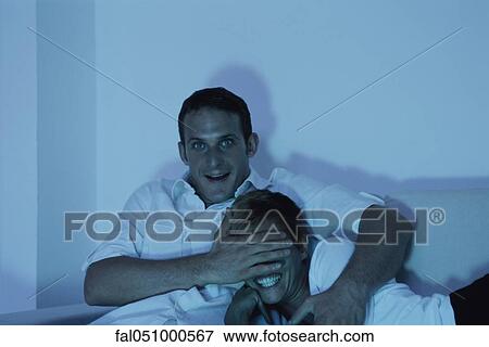 Couple watching TV together, man covering woman's eyes with his hand View Large Photo Image Stock Photo - Couple watching TV together, man covering woman's eyes with his hand. Fotosearch