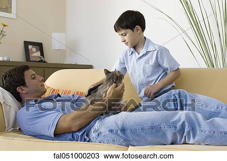 Stock Image - Man relaxing with cat on lap, young son brushing cat. Fotosearch