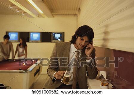 Stock Image - Man talking on cell phone in bar with glass of beer in hand, people playing pool in background. Fotosearch