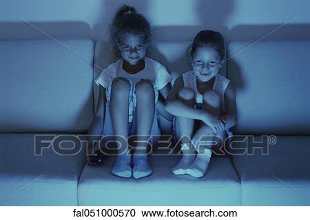 Stock Image - Two girls watching TV together at night, both smiling. Fotosearch