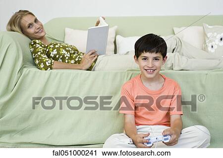 Young boy sitting on floor playing video game, mother relaxing on sofa View Large Photo Image Stock Image - Young boy sitting on floor playing video game, mother relaxing on sofa. Fotosearch