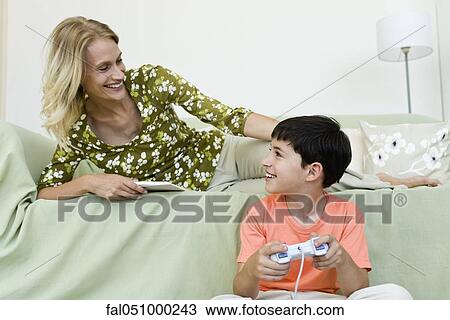 Young boy sitting on floor playing video game looking up at mother relaxing on sofa View Large Photo Image Stock Image - Young boy sitting on floor playing video game looking up at mother relaxing on sofa. Fotosearch