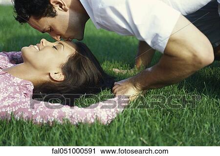Stock Image - Young couple relaxing on grass, man kissing woman's forehead. Fotosearch